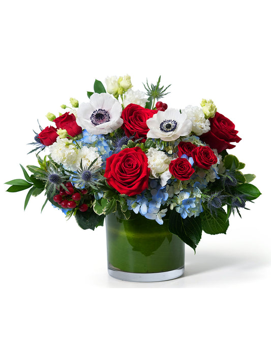 Vibrant bouquet of red roses, white anemones, and blue hydrangeas in a glass vase, perfect for any occasion.